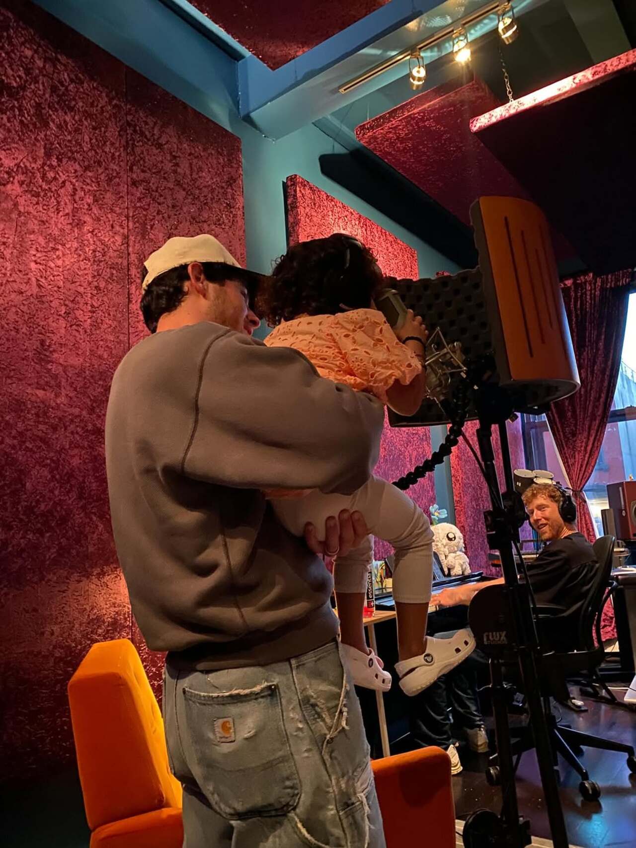 In this heart-melting moment, Nick Jonas is seen holding his daughter Malti in a vibrant recording studio. Nick lovingly supports Malti as she leans toward the microphone with curiosity and excitement.